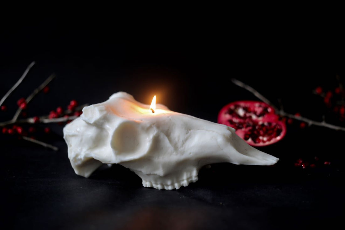 Evangeline Linens soy Deer Skull Candle lit on a moody black background. A bright red halved pomegranate sits behind the candle.