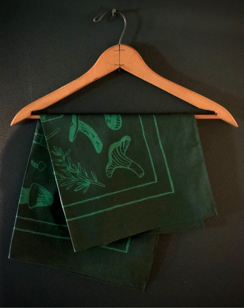 Green and green evangeline bandana folded and hanging on a hanger. Bandana has illustrations of mushrooms and lavender.