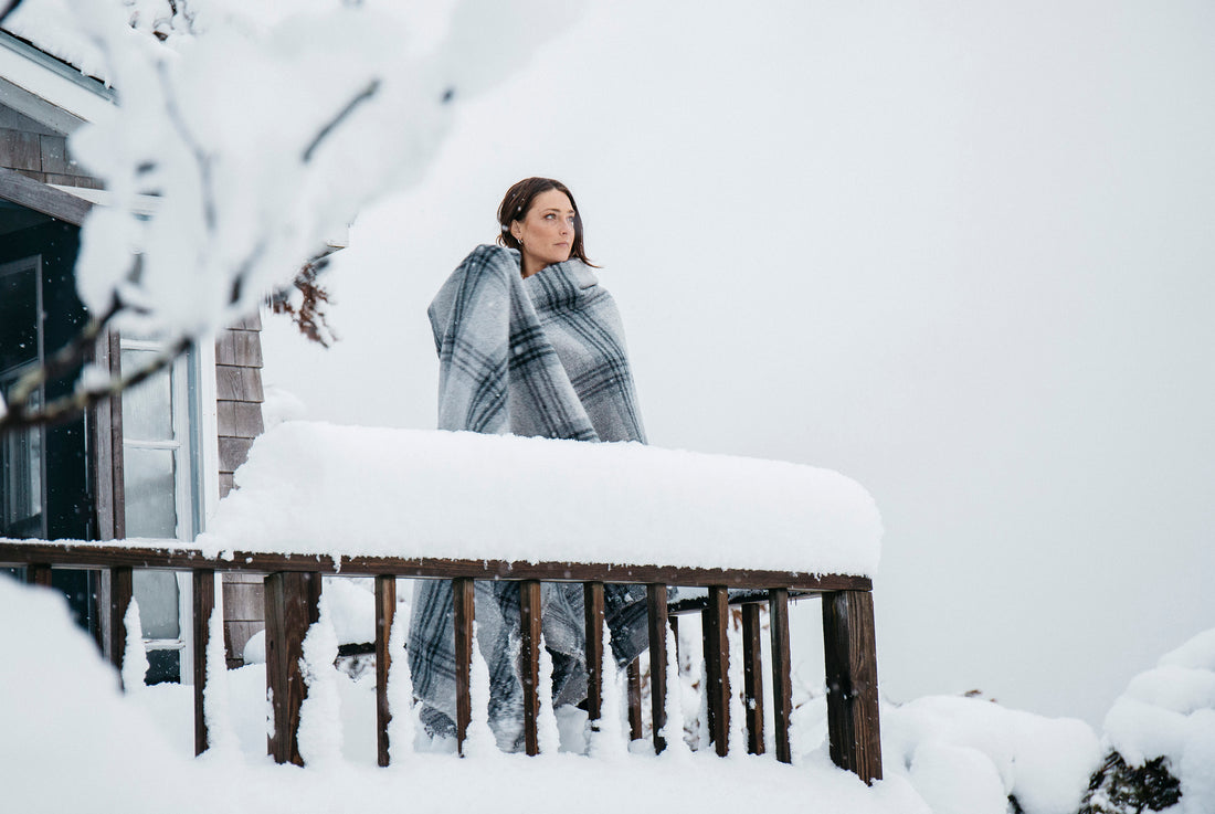 Woman wrapped in Evangeline Linens plaid merino wool bed blanket on a snowy porch.