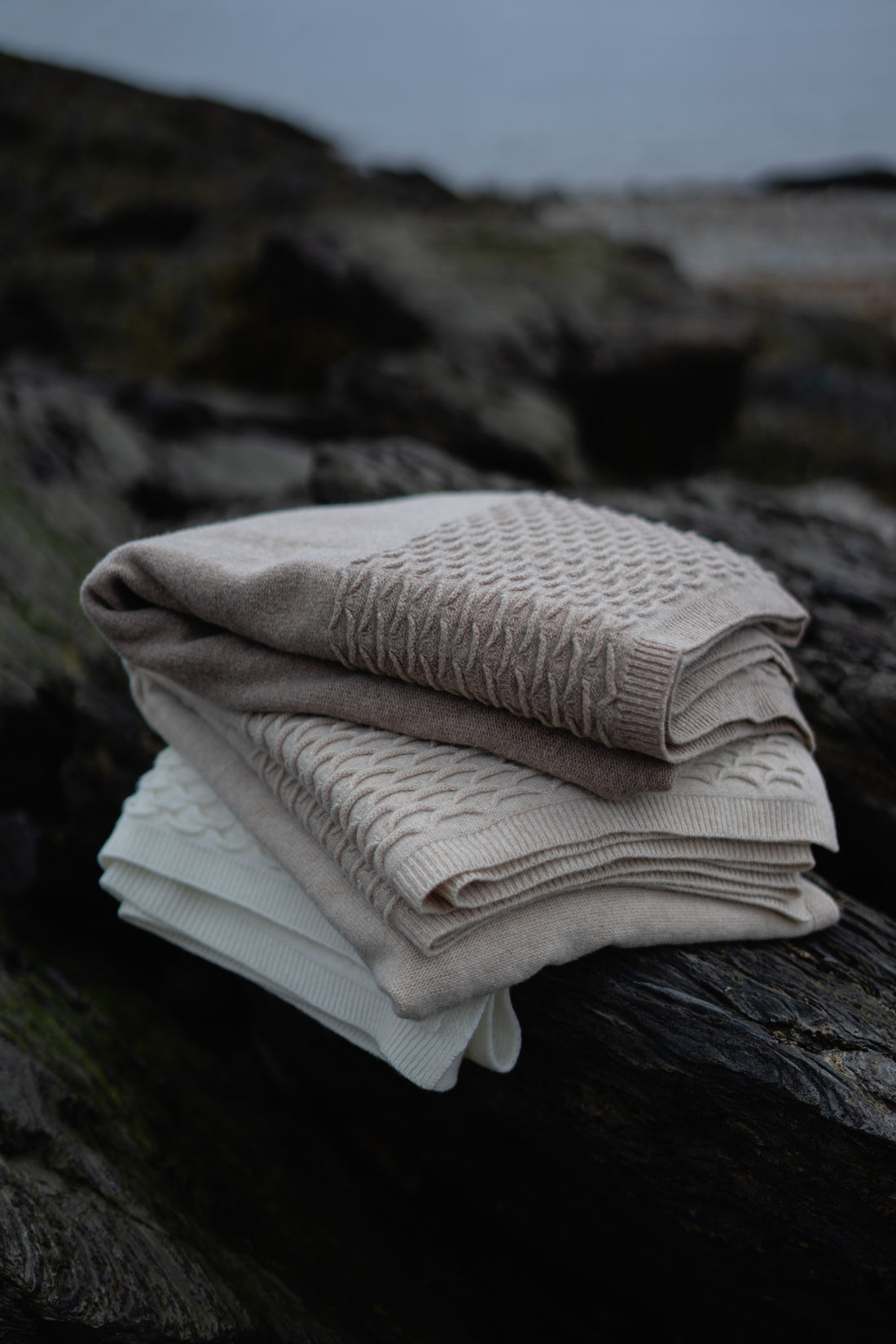 A folded stack of Evangeline Knit 100% soft lambswool throws, featuring a scalloped detail. Stack includes Coffee, Oatmeal, and Pearl (top to bottom). Super soft 'hand' of lambswool. Unique scallop texture. Has the energy of a treasured knit sweater.