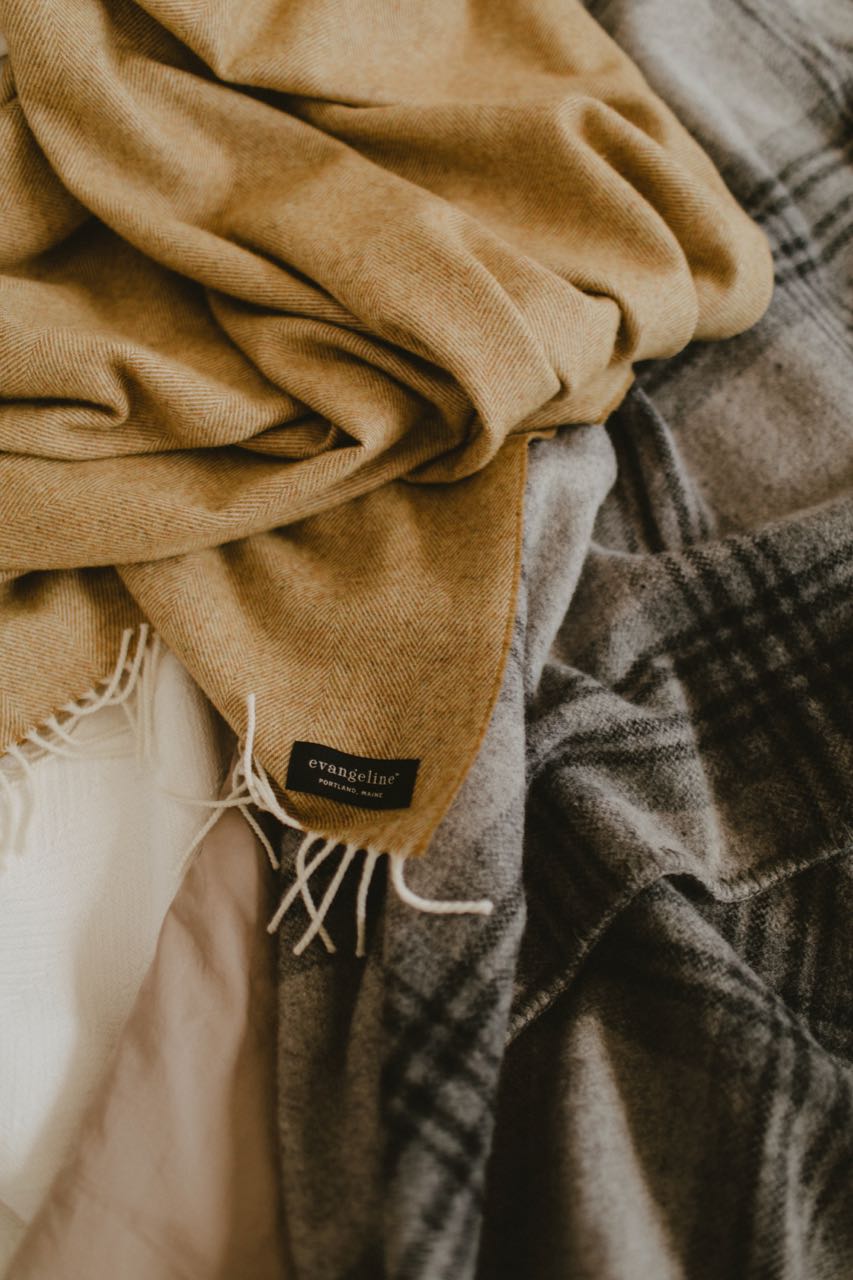 A detail shot of Evangeline's lightweight wool herringbone throw and the sewn Evangeline tag. Lays on top of an Evangeline wool bed blanket on a bed. The lighter weight 'sibling' to our popular Herringbone Throws.  Oh-so-soft. And perfect for summer nights.  