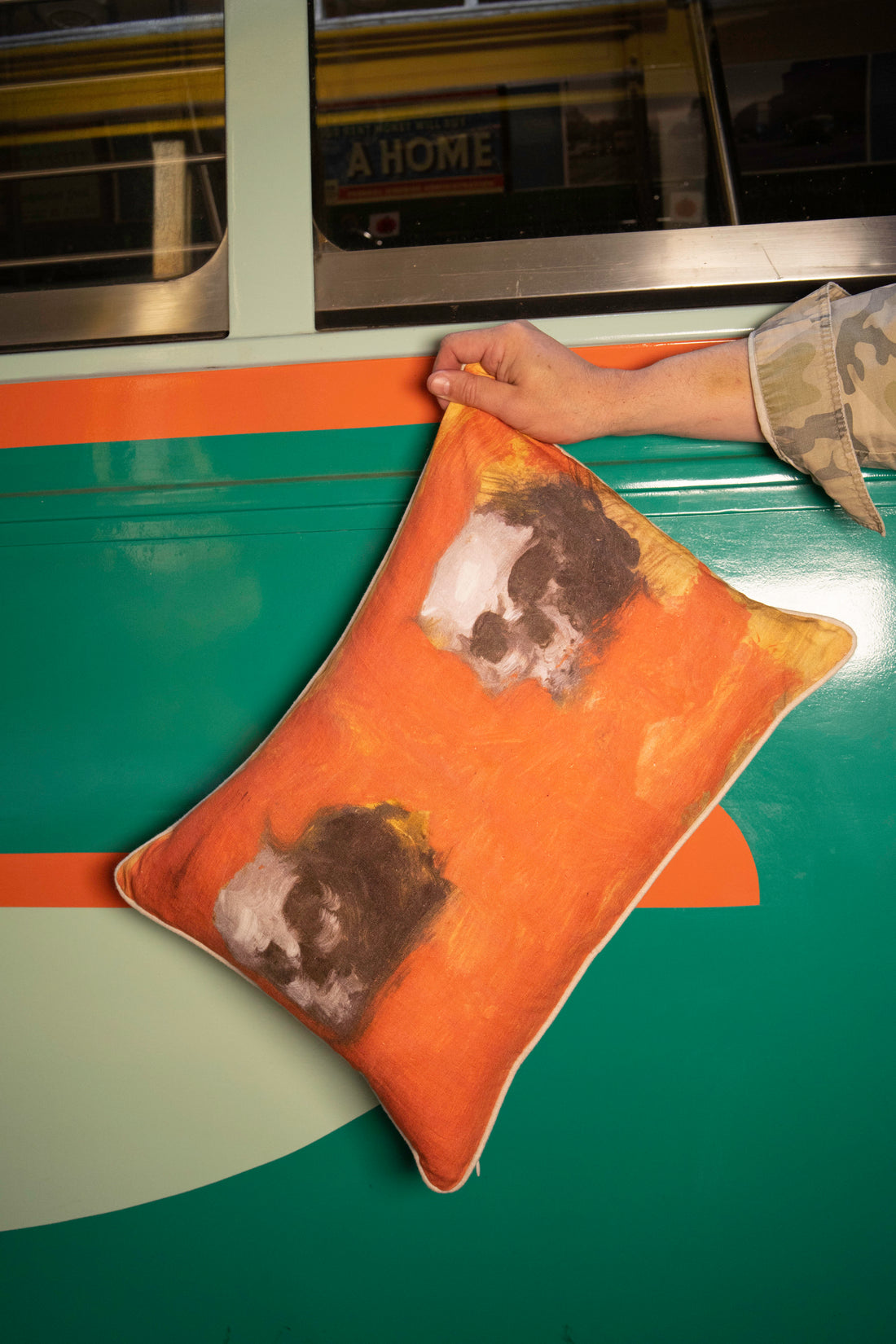 A linen pillow with a painted artwork printed on it, depicting two human skulls against an orange background, is held by a persons arm in front of the side of a trolley. The 100% Natural European Linen Pillow Includes 10/90 Down / Feather Blend Insert. Sophisticated and refined. Modern yet classic. A true friend for years to come. Dimensions: 17"x24" 