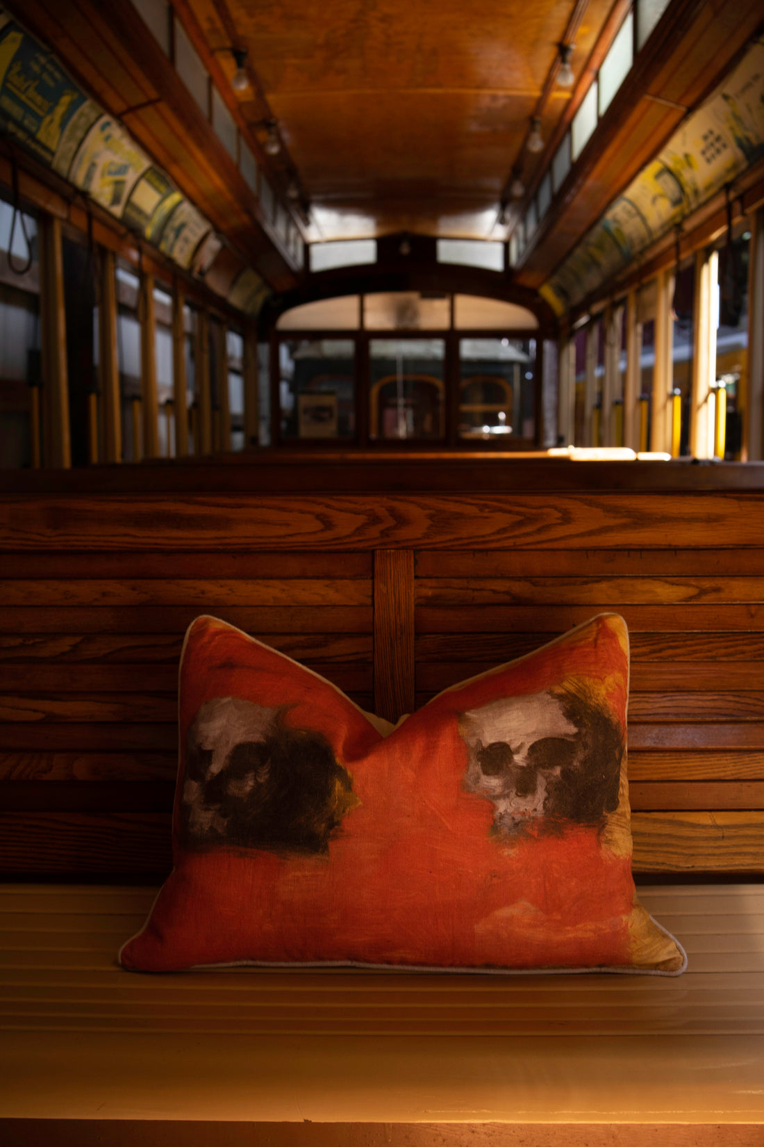 Evangeline's linen 'Twins' pillow, orange with two human skull images, sits on a wooden bench in vintage trolley.