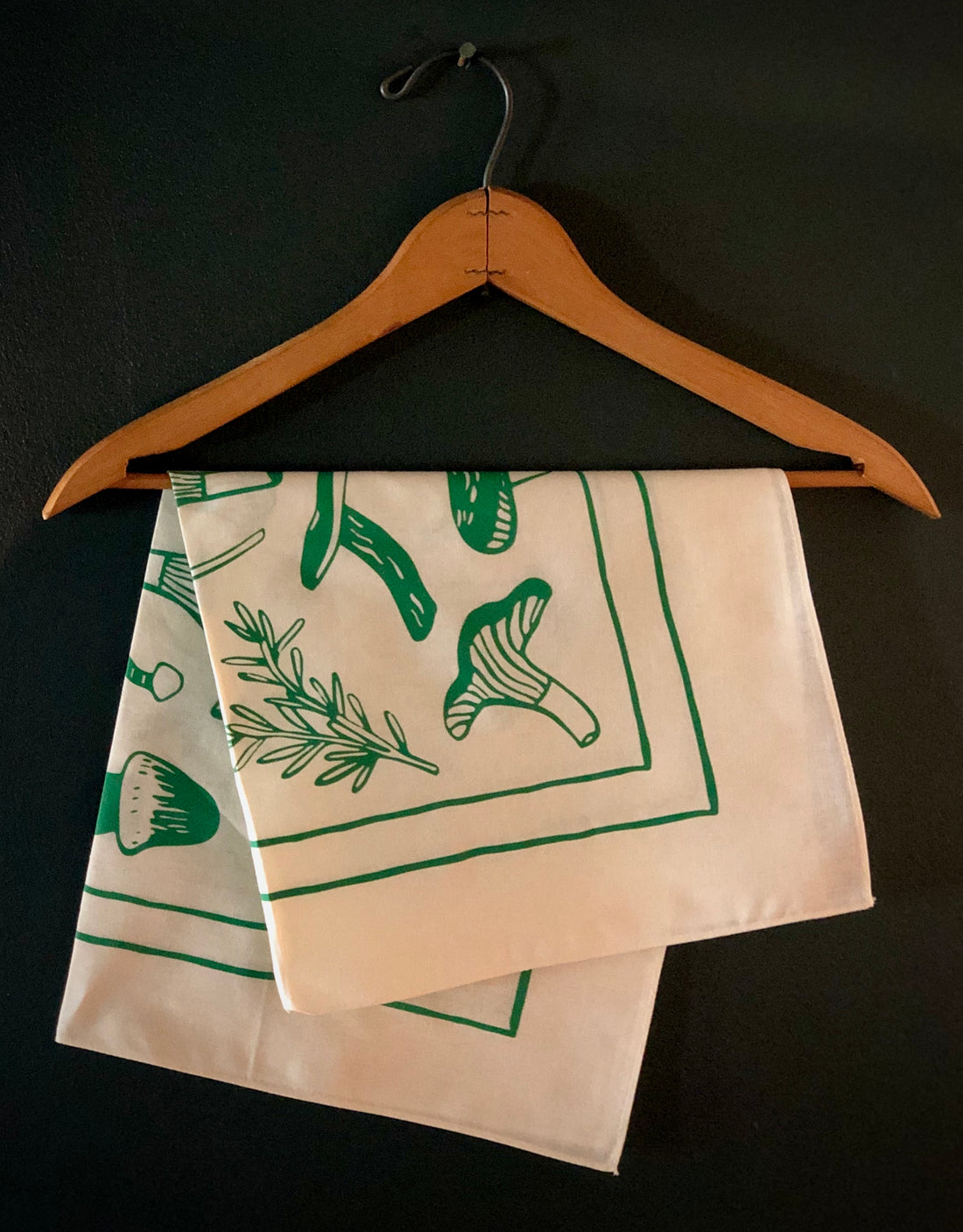 Cream and green evangeline bandana folded and hanging on a hanger. Bandana has illustrations of lavender, rosemary and mushrooms.