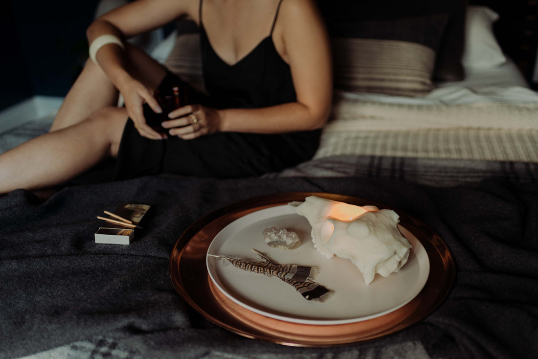 Evangeline Linens soy Deer Skull Candle lit on a tray placed on a made bed. A woman with medium skin tones sits on the bed behind the candles just out of focus.