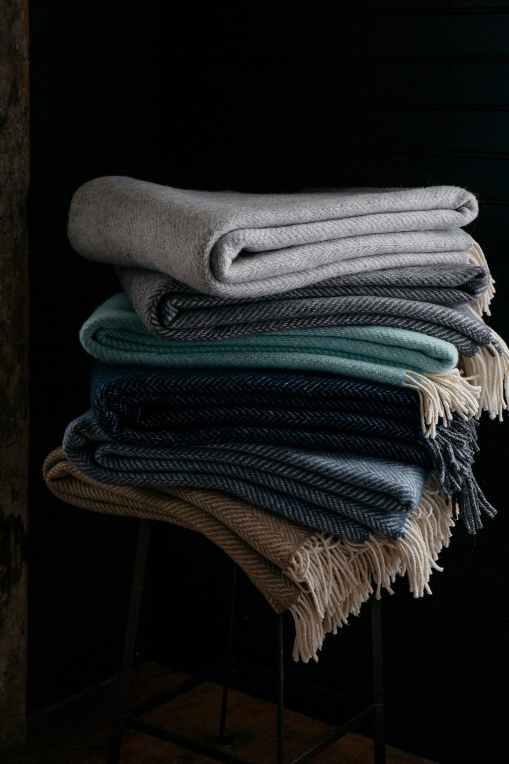 A stack of folded Merino Lambswool Cashmere throws. A beautiful display of Six out of 10 different colorways. Light grey fog, darker grey graphite, turquoise sea foam, deep midnight blue with a grey fringe detail, denim blue twilight and finally our beige colored harvest color herringbone throw.