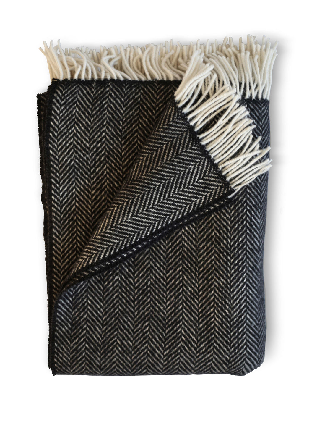 Folded Herringbone throw in Thundercloud, a dark grey colored throw with cream tassel detail. Timeless herringbone design. Super soft luster of cashmere. cozy comfort of merino wool. A chunky and cuddly classic. 95% Merino Lambswool and 5% Cashmere. Woven in Ireland.