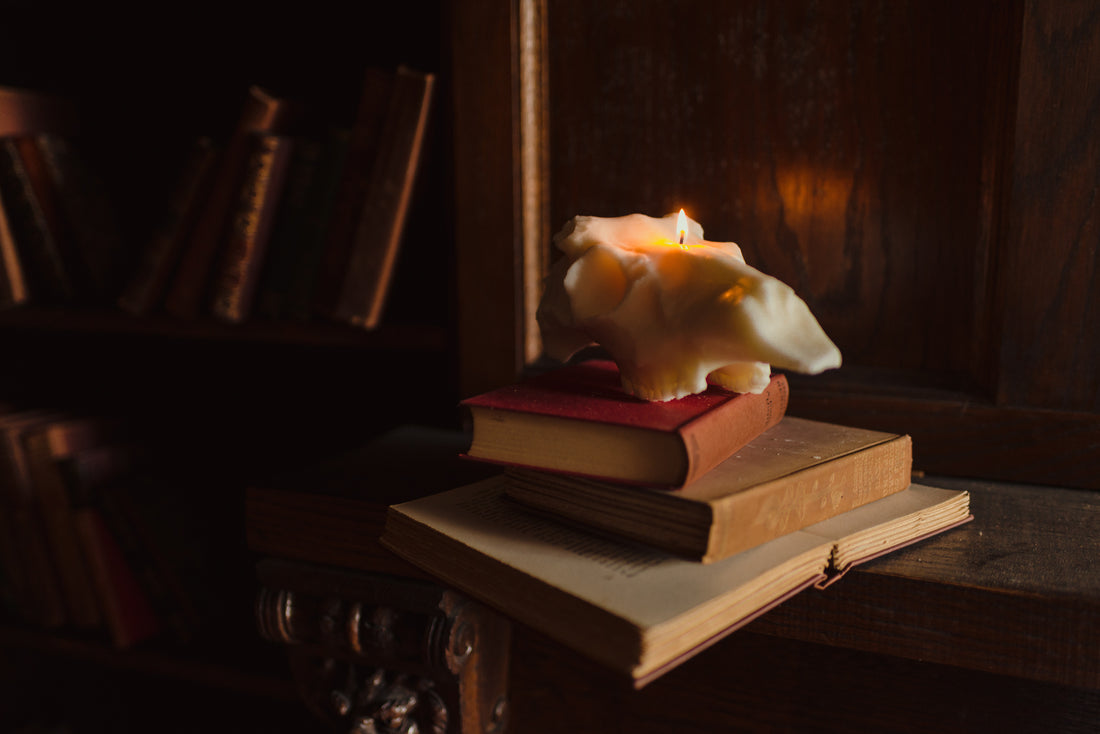 Evangeline Linens soy Deer Skull Candle lit on a stack of books placed on a mantle in an old library. Skull candles are made of soy wax and are hand-poured in Portland Maine in small batches.
