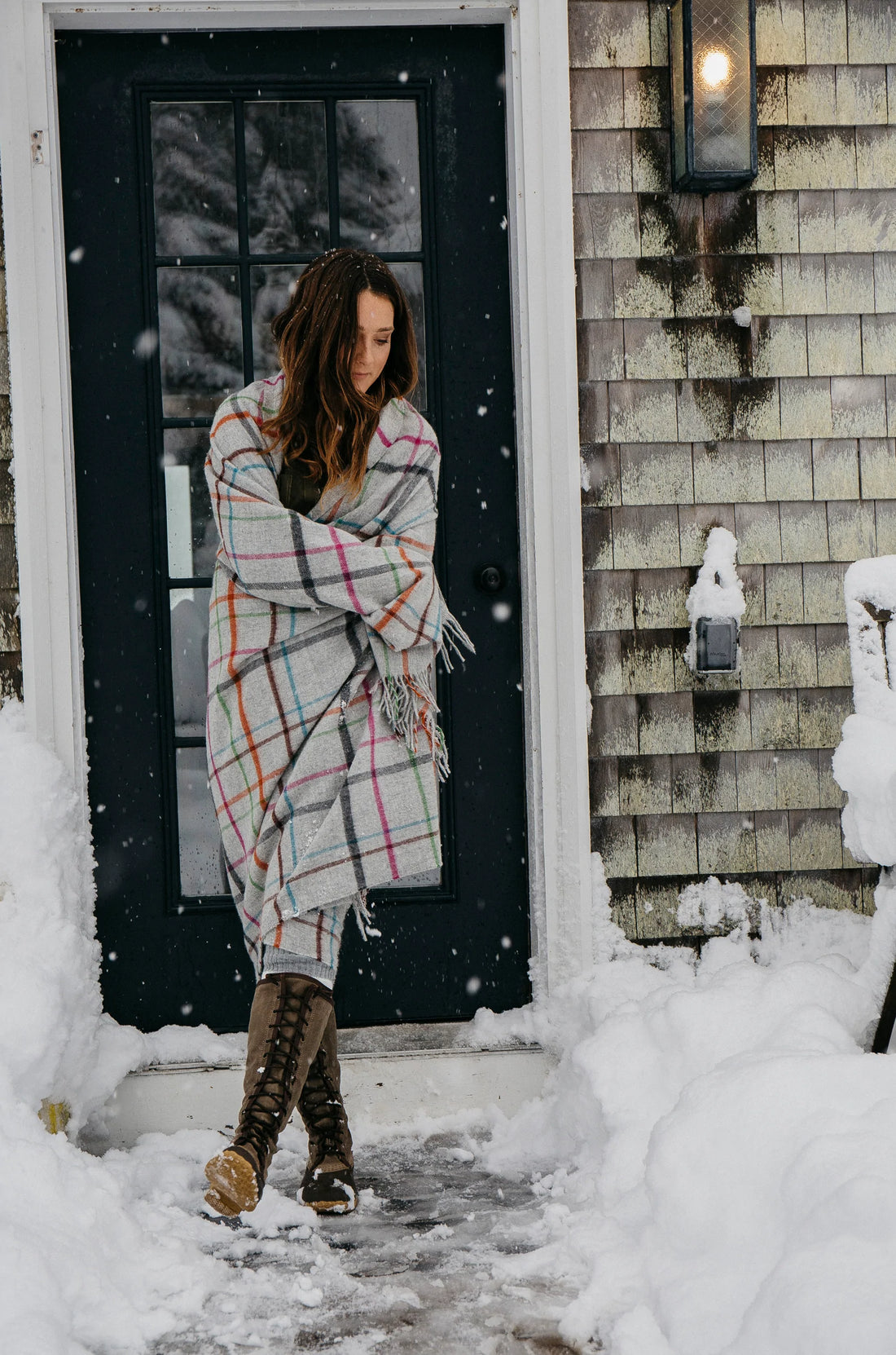 Woman stands in a snowy scene with merino lambswool light grey multi windowpane throw blanket around her shoulders. 