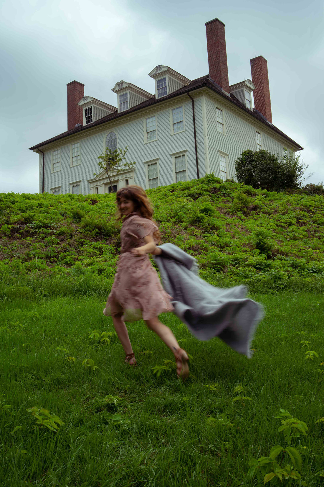 A woman running with a dawn / light blue / mohair throw blanket. A historic house sit up on a hill in the distance. 