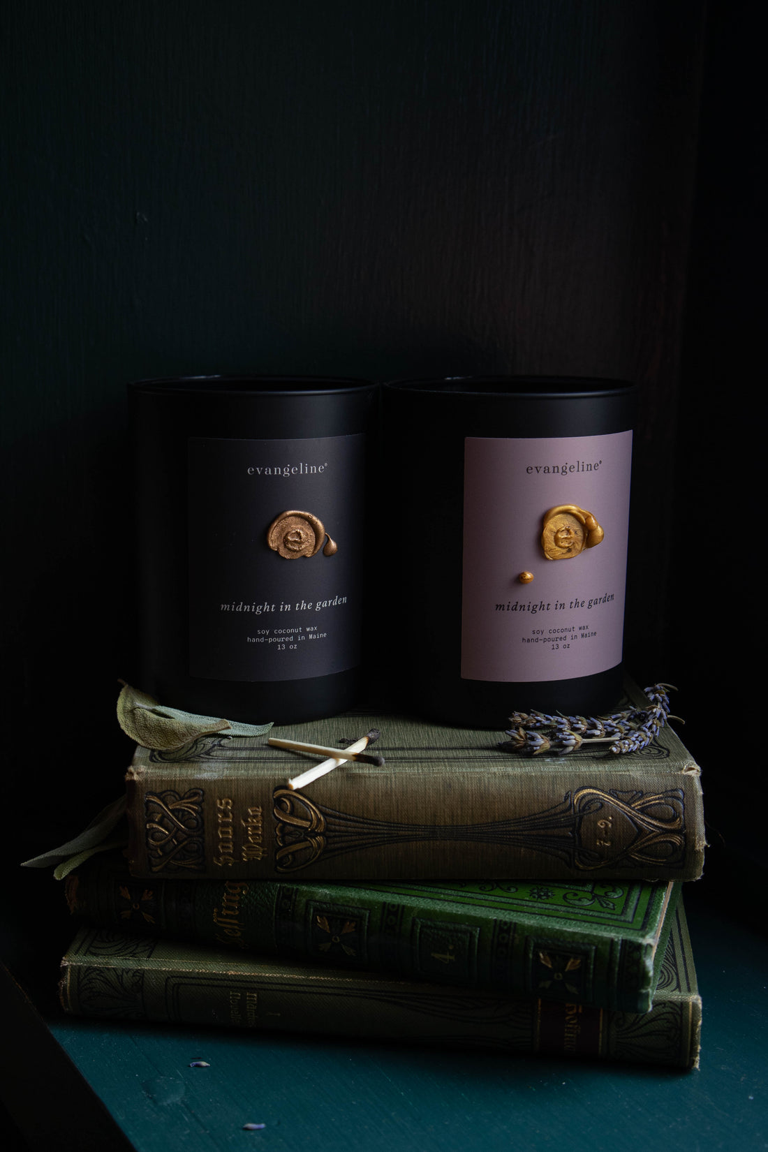 Two Matte black glass candles with matte black and blush midnight in the garden labels, hand-poured gold wax seal, surrounded by lavender, sage , moss and oak.
