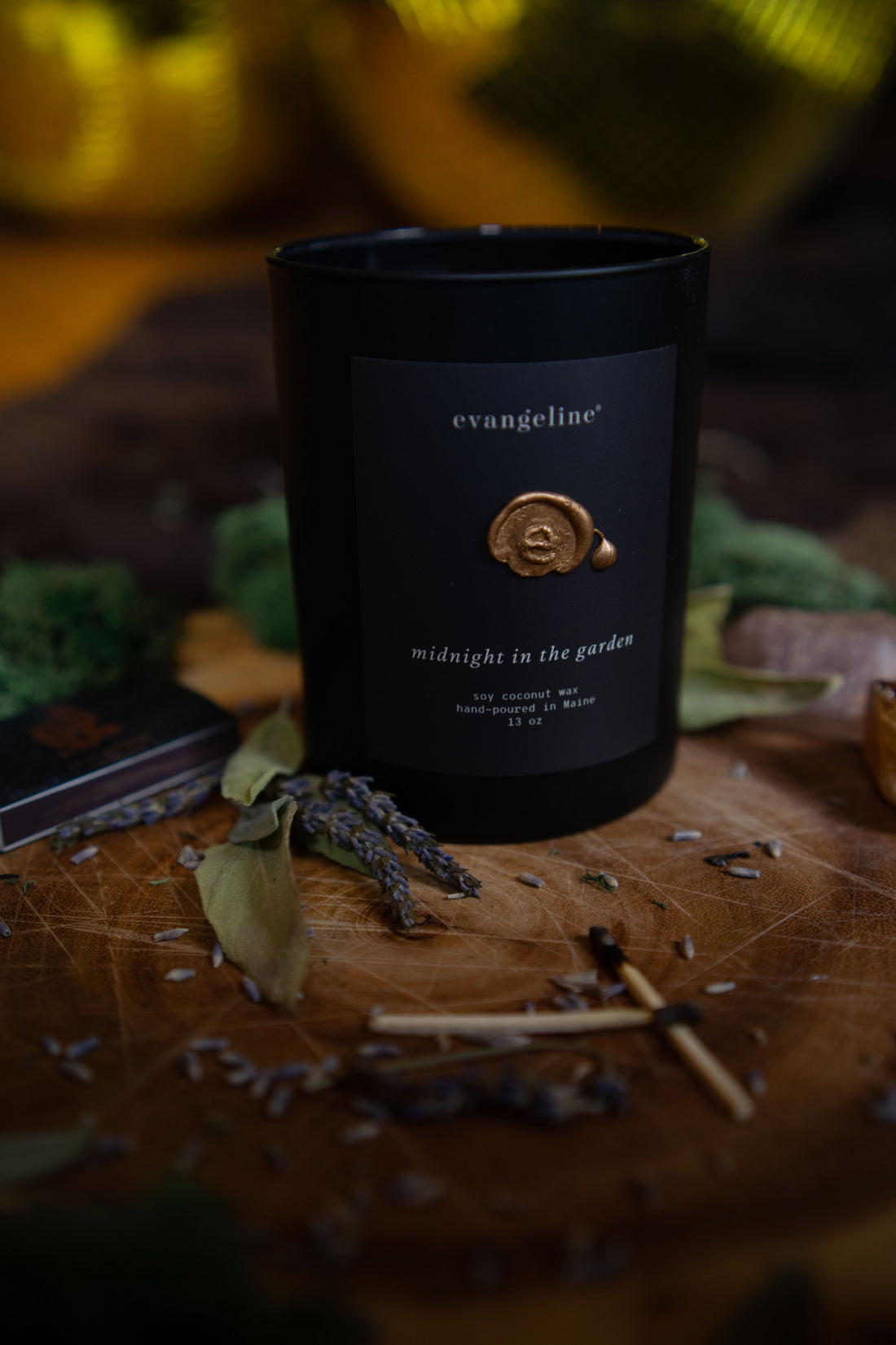 Matte black glass candle with matt black midnight in the garden label, hand-poured gold wax seal, surrounded by lavender, sage , moss and oak.